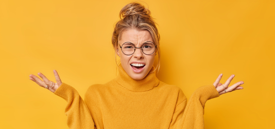 Junge Frau in gelbem Pullover steht vor gelbem Hintergrund und schaut fragend in die Kamera, mit hochgezogenen Augenbrauen und offenen Händen.