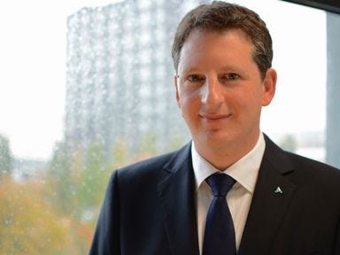 Mann mit dunklem Anzug und Krawatte steht vor einem Fenster und l&auml;chelt