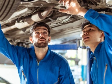 Zwei junge Männer in blauer Arbeitskleidung arbeiten gemeinsam an einem Auto in einer Werkstatt.