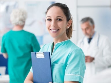 Junge Pflegekraft in grüner Kleidung hält ein Klemmbrett, im Hintergrund ältere Menschen und Kolleg:innen.