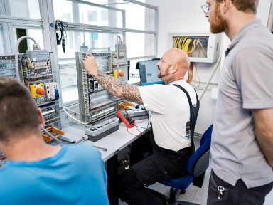 Zwei Teilnehmer sitzen vor einer Elelktrotechnischen ANlage hinter ihnen steht der Ausbilder 