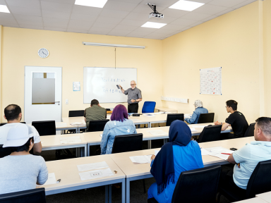 Mehrere Teilnehmer sitzen in einem Sprachkurs und der Dozent steht vorne an einem Whiteboard