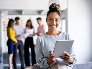 Qualifizierungsgeld – Mehr Geld für Weiterbildung Eine Frau steht mit einem Tablet im Vordergrund in einem Büro und im Hintergrund stehen ihre Kollegen