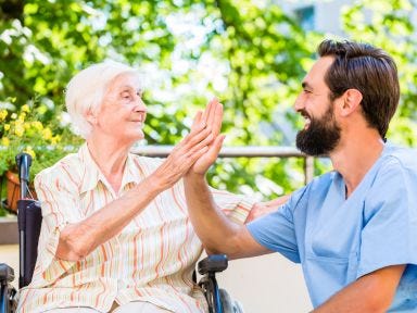 Ein m&auml;nnlicher Pfleger geht mit einer &auml;lteren Dame in einem Rollstuhl spazieren