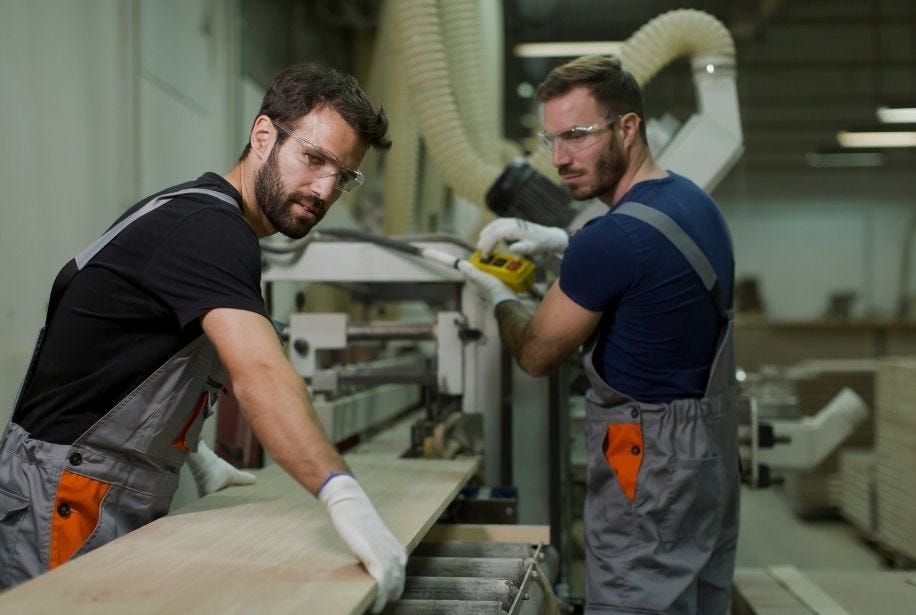 Zwei M&auml;nner mit Schutzbrille arbeiten an einer Maschine in der Holzwerkstatt.