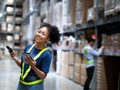 Ein Mitarbeiterin hält im ein Tablet in der Hand und steht in einer großen Lagerhalle und ein Mitarbeiter steht an einem Regal