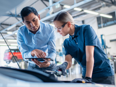 Ausbilder erklärt einer Auszubildenden in der Kfz-Werkstatt technische Details am Fahrzeug.