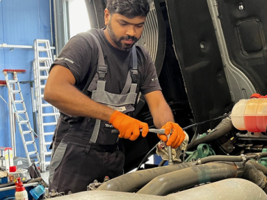 Junger Kfz-Mechatroniker mit orangefarbenen Handschuhen arbeitet konzentriert im Motorraum eines Fahrzeugs.