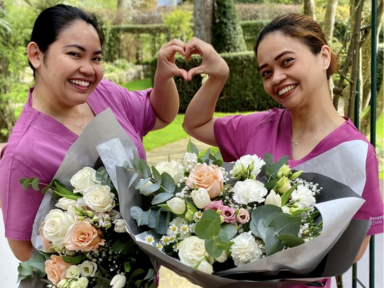 Internationale Pflegekräfte der Rosenpark Klinik Zwei junge Pflegekräfte in lilafarbener Kleidung lächeln und formen gemeinsam ein Herz mit ihren Händen, während sie einen großen Blumenstrauß halten.