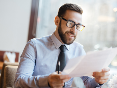 Philipp Aloe über die Ausbildung bei TÜV Rheinland Geschäftsmann mit Brille liest lächelnd ein Dokument in einem hellen Büro – symbolisiert Zufriedenheit mit Ausbildungspartnern.