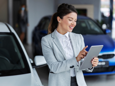 Eine junge Frau in Business-Kleidung steht mit einem Tablet in einem Autohaus oder Büro.