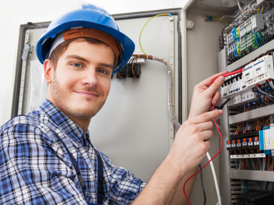 Ein junger Mann mit Schutzhelm arbeitet lächelnd an einem Schaltschrank mit elektrischen Leitungen.