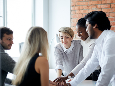 Teamarbeit in der Ausbildung mit TÜV Rheinland Fünf junge Menschen verschiedener Herkunft in einem modernen Büro, die sich gegenseitig motivierend die Hände reichen – symbolisiert Teamwork und Zusammenhalt